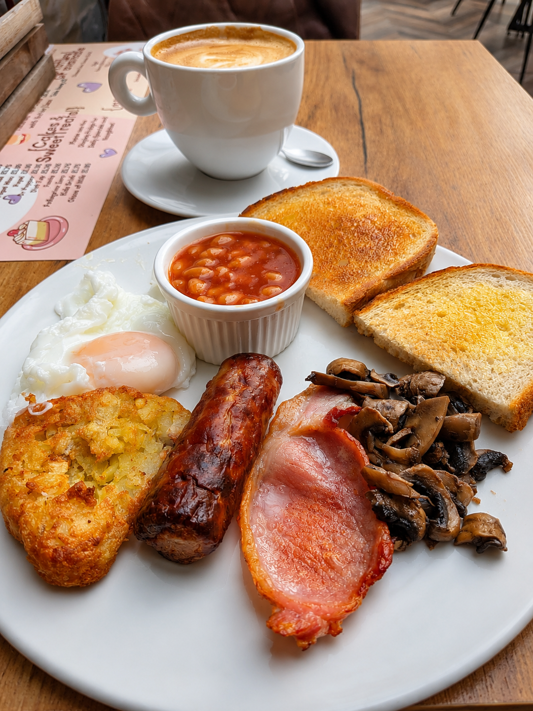 A full breakfast plate with eggs, bacon, sausage, mushrooms and toast beside coffee